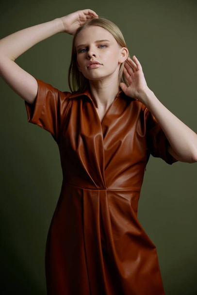 Výrazná elegantní mladá sebevědomá blondýnka v módním stylovém koženém hnědém plášti, který představuje izolované dojemné vlasy držící ruce nad hlavou na pozadí studio zelené stěny. Módní banner nabídka - Fotografie, Obrázek