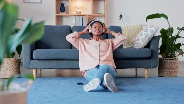 Woman listening to music and enjoying songs on wireless headphones, sitting on the living room floor inside. Young trendy african female with afro relaxing to her favorite podcast and meditating. - Video