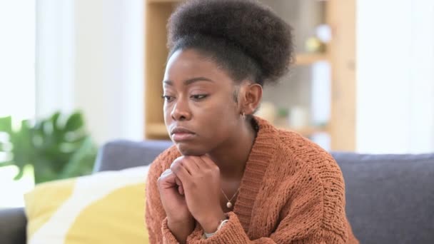 Smutna Afroamerykanka siedząca sama na kanapie w domu. Portret zmęczonej kobiety sfrustrowanej i nieszczęśliwej. Zdenerwowana kobieta wyglądająca na zestresowaną i przygnębioną problemami z pracą na kanapie - Materiał filmowy, wideo