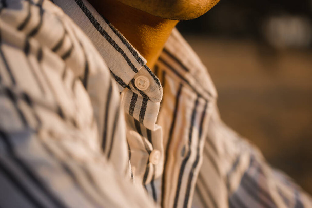 Faceless dark-skinned man, a young guy in a white blue striped shirt. Detail of business men's clothing. Adult male relaxing in nature, taking a break from work. Emphasis on shirt collar, buttons. - Zdjęcie, obraz