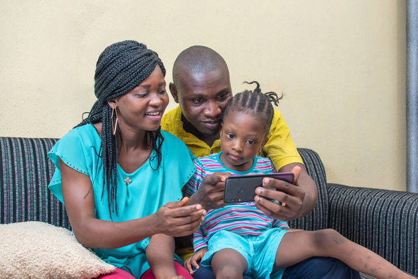 Una famiglia di tre membri rannicchiati e seduti insieme su un divano con uno smartphone in mano, il gruppo è composto da una bambina africana, una donna e un uomo. - Foto, immagini