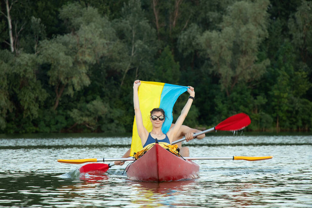 Uma menina e um cara estão navegando em uma canoa ao longo do rio Dnieper, na cidade de Dnepr, na Ucrânia, ao pôr do sol, com a bandeira da Ucrânia, durante a guerra nos dias de bombardeio da cidade 2022. - Foto, Imagem