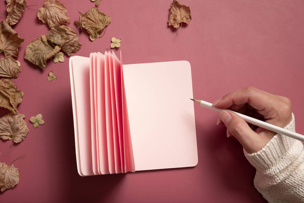 Table desk with blank pink paper note pad and female hand wit pencil and fall leaves on colored background. Autumn flat lay composition with blank paper. - Foto, Imagem