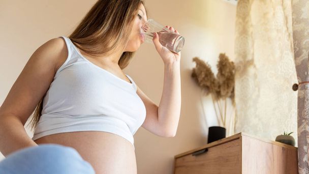 Ciąża pije wodę. Piękna ciąża pijąca wodę. Szczęśliwa ciężarna kobieta trzymająca szklankę wody. Ciąża, macierzyństwo, koncepcja opieki zdrowotnej - Zdjęcie, obraz
