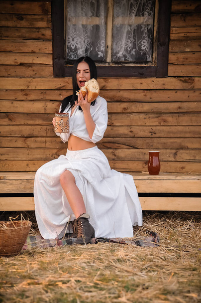 mooie jonge vrouw in een witte jurk en strohoed zittend op een houten bank in een zomerdag - Foto, afbeelding