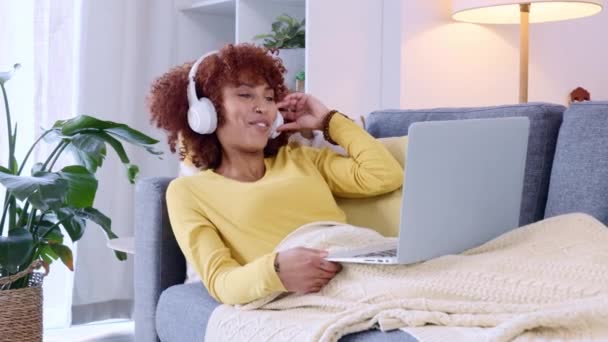 Woman on a video call, talking to friends or family and relaxing on the sofa at home, looking happy and relaxed. Trendy young female chatting on a laptop and wireless headphones online on the couch. - Imágenes, Vídeo