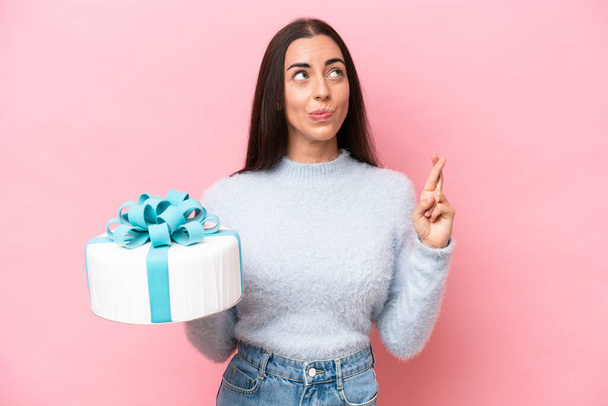 Young caucasian woman holding birthday cake isolated on pink background with fingers crossing and wishing the best - Photo, image