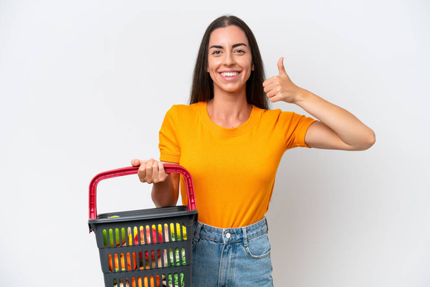 Young caucasian woman holding a shopping basket full of food isolated on white background giving a thumbs up gesture - 写真・画像