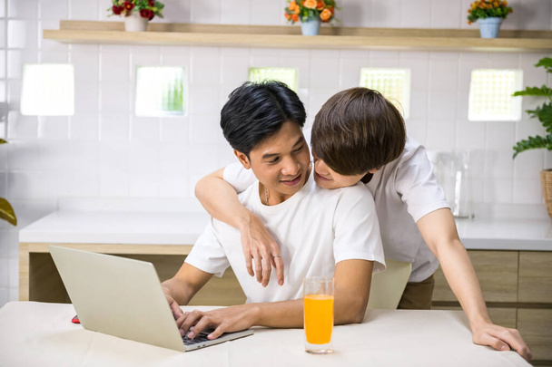 A fiatal LMBT ázsiai meleg férfi megöleli a társát, aki a laptop számítógépén dolgozik hátulról. Boldog pár együtt otthon. Ugyanaz a család.. - Fotó, kép