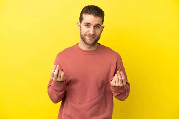 Handsome blonde man over isolated yellow background making money gesture - Foto, afbeelding