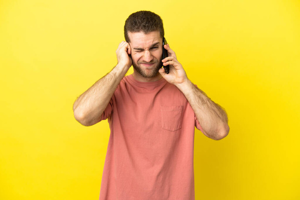 Handsome blonde man using mobile phone over isolated background frustrated and covering ears - Zdjęcie, obraz