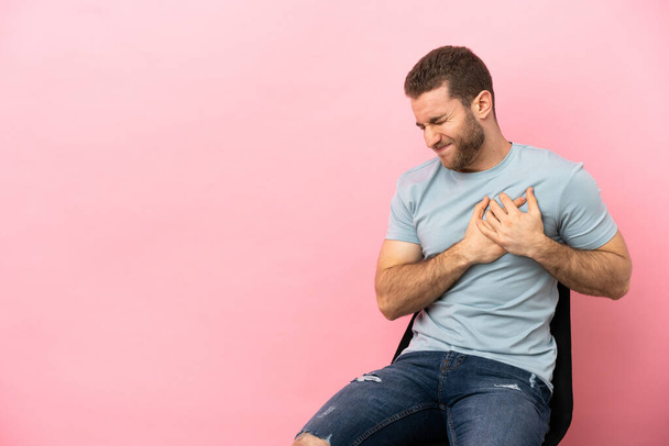 Young man sitting on a chair over isolated pink background having a pain in the heart - Foto, Bild