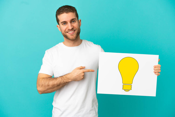 Schöner blonder Mann über isoliertem blauem Hintergrund, der ein Plakat mit einem Lampensymbol hält und darauf zeigt - Foto, Bild