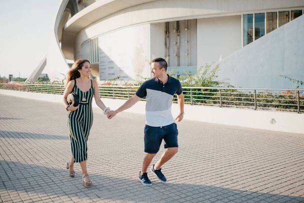 A romantic guy is holding his smiling girlfriend in the modern urban space at the sunset in a Spain town. A couple of tourists is running in the evening in Valencia. - Φωτογραφία, εικόνα