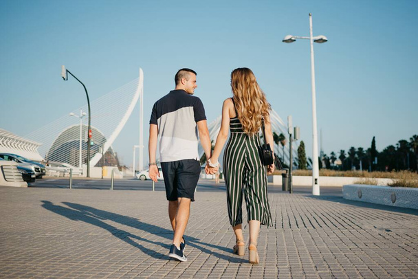 A photo from behind a couple of tourists is walking in the modern urban space at the sunset in Valencia. A romantic guy is holding hands with his girlfriend in the evening in Spain. - Foto, Imagem