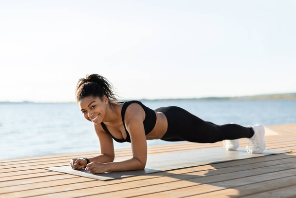 Cheerful attractive young african american woman doing workout at seaside, planking on fitness mat and smiling at camera, athletic lady working on body strength, copy space - Foto, imagen
