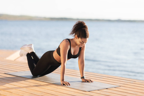 Athletic young black woman in black tight sportswear exercising by water river or lake at public park, stretching on fitness mat, practicing morning yoga outdoors, enjoying sunny day, copy space - Φωτογραφία, εικόνα