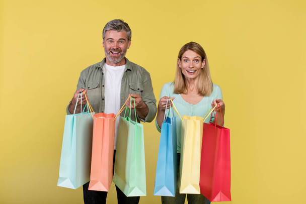 Big sale and shopping with pleasure. Excited middle aged spouses holding many colored packages in hands, posing on yellow background, studio shot, copy space - Fotó, kép