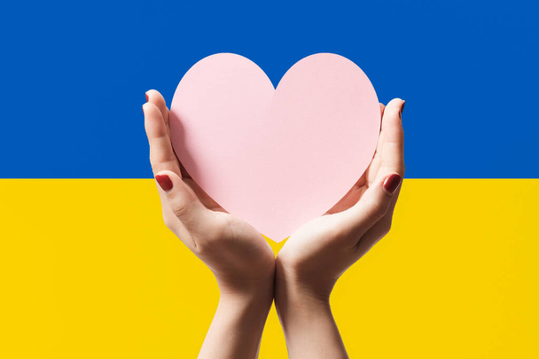 Donne mani in possesso di carta a forma di cuore su sfondo di colore della bandiera Ucraina. Resta con il simbolo ucraino. Mano cuore amore gesto con sfondo bandiera ucraina. - Foto, immagini