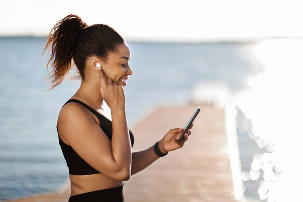 Sportliche schwarze Millennial-Frau, die auf Ohrhörer drückt, auf Smartphone-Bildschirm schaut und lächelt, positive athletische Dame, die Musik zum Workout einschaltet, Training am See im öffentlichen Park, Kopierraum - Foto, Bild