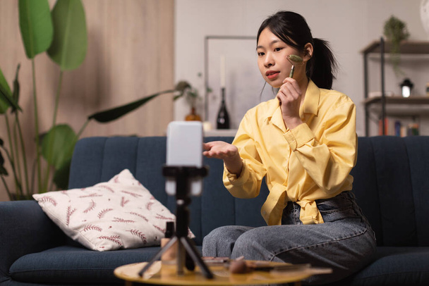 Koreanisch blogger dame herstellung ins gesicht massage mit stein roller und filmen video auf mobile phone für blog sitting on couch at home. Professionelles Beauty Blogging. Selektiver Fokus - Foto, Bild
