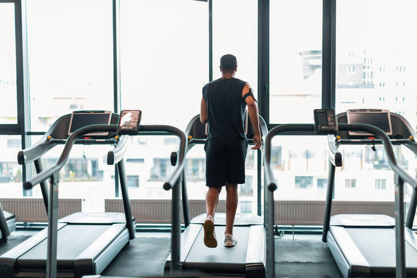 Sporty Lifestyle. Unrecognizable Black Man Jogging On Treadmill Near Panoramic Window At Gym, Rear View Of Young African American Athlete Training On Modern Equipment At Sport Club, Copy Space - 写真・画像