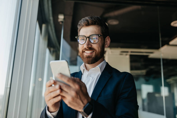 Szczęśliwy odnoszący sukcesy biznesmen w okularach piszący na smartfonie w nowoczesnym wnętrzu biurowym, mający czat z klientem online. Biznes w miejscu pracy, przerwa w pracy - Zdjęcie, obraz