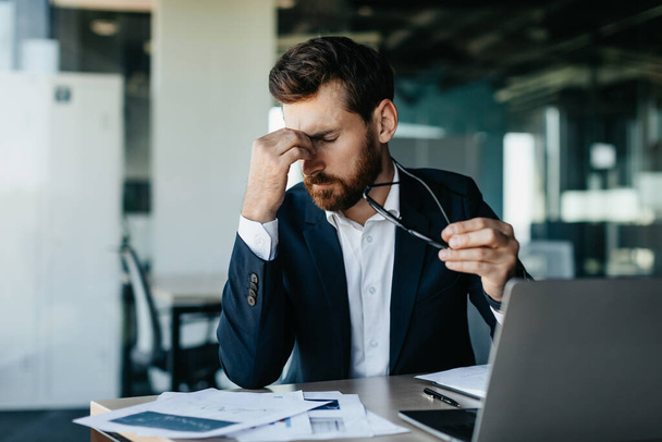 Exhausted male manager or businessman sitting in front of laptop at modern office interior, holding glasses and rubbing nose bridge, having headache - Фото, изображение