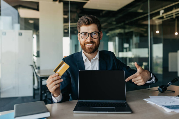 Happy businessman holding credit card and pointing at laptop screen with empty screen, sitting in office, mockup. Male entrepreneur purchasing and paying in internet store - Foto, imagen