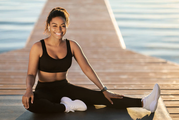 Happy pretty young african american woman in tight black sportswear posing while exercising outdoor next to city lake, sitting on fitness mat and smiling at camera, copy space - Fotó, kép