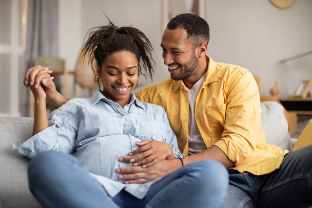 Black Millennial Couple Embracing Expecting Baby, Boyfriend Touching Girlfriends Belly Sitting On Sofa In Living Room At Home. Pregnancy Happiness And Lifestyle, Childbirth Concept - Foto, Bild