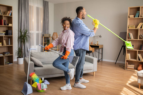 Happy African American Couple Doing Housework Cleaning House Together And Singing Having Fun Posing With Broom And Mop Standing In Modern Living Room At Home. Full Length - Foto, imagen