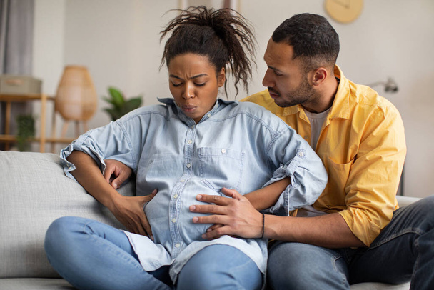 Terhes afro-amerikai nő szenved a munkásfájdalomtól, a férj ölelkezik, támogatja őt otthon a kanapén. Feleség szülés a házban. Terhesség és szállítás koncepció - Fotó, kép