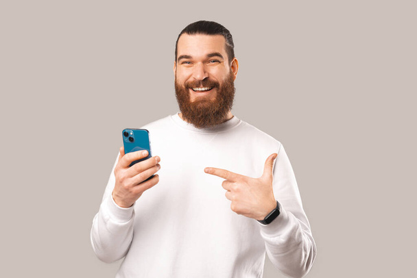 A photo of a bearded man pointing to his phone and while looking at the camera he is smiling - Fotografie, Obrázek