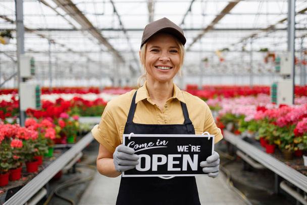 Pretty woman owner of a flower greenhouse with a sign come in we're open - Photo, Image