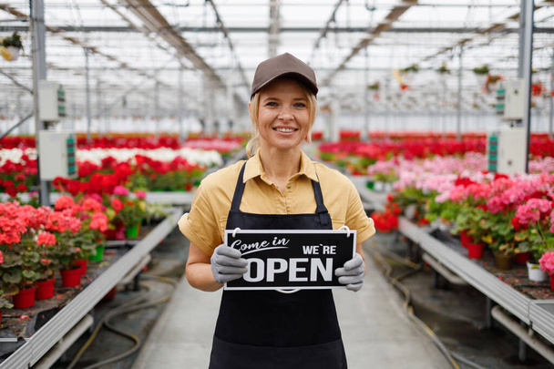 Portrait of pretty female flower greenhouse worker showing sign we are open - Foto, immagini