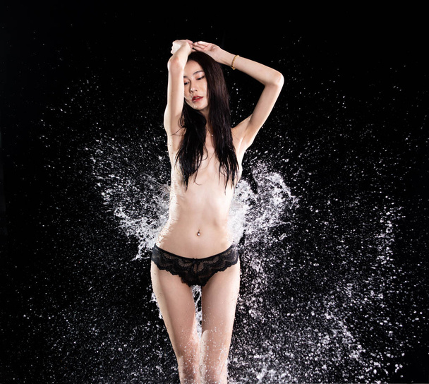 Young 20s Asian Woman wear topless black lace lingeries with water splash throw to back of model, long black hair over black background. Female poses sexy sensual fashion vintage water splashing - Foto, imagen