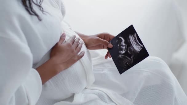 Amor de maternidade. Close-up tiro de mulher grávida afro-americana irreconhecível admirando imagem de ultrassonografia de seu bebê por nascer e acariciando barriga, sentado em casa, câmera lenta - Filmagem, Vídeo
