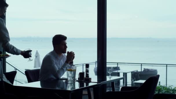 Elegant waiter silhouette serving successful businessman customer in seascape restaurant. Handsome man resting weekend lunch in lounge hotel bar. Cafe at covid 19 concept. Luxury lifestyle concept. - Materiaali, video
