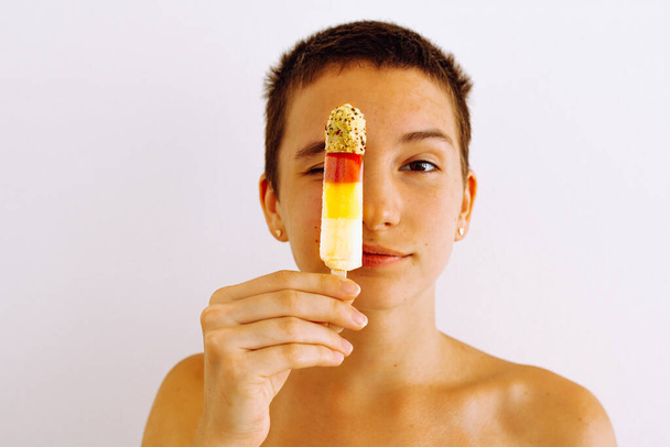 Caucasian teenage girl hides behind popsicles, attractive girl with short hair, bare shoulders, shows emotions, pursed lips, in playful manner of young woman. Attractive teenager shows frozen fruit - Foto, Bild
