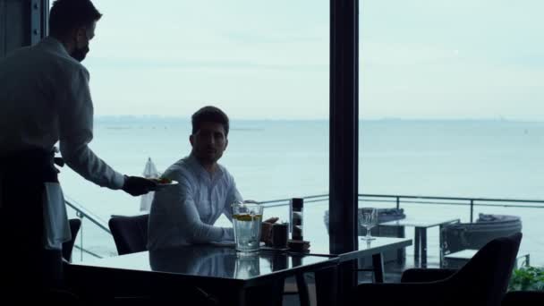 Relaxed man resting hotel cafe terrace at lunch time break. Elegant masked waiter serving meal to successful businessman customer. Joyful entrepreneur silhouette enjoying sea view in luxury restaurant - Video