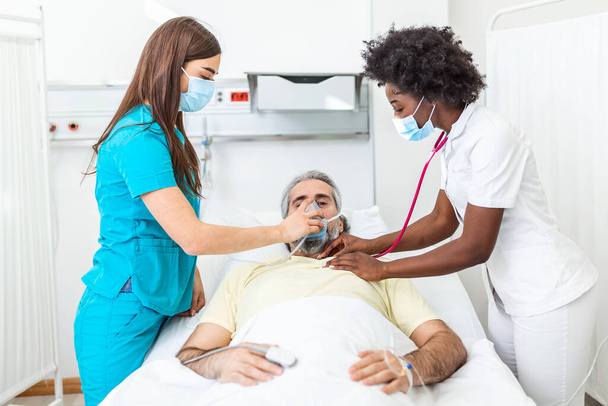 Nurse puts oxygen mask on elderly man patient lying in the hospital room bed while African American doctor examining patient with stethoscope, coronavirus covid 19 protection concept - Photo, image