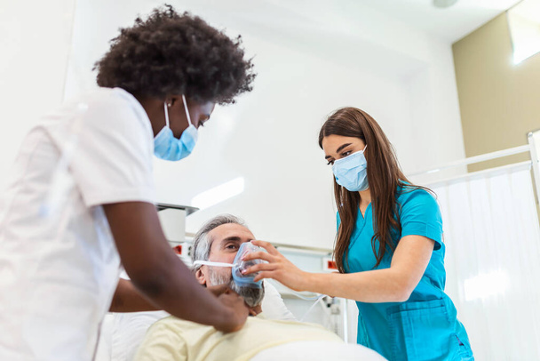 Krankenschwester legt Sauerstoffmaske auf älteren Mann im Krankenhausbett liegend, während afroamerikanischer Arzt Patient mit Stethoskop untersucht, Coronavirus covid 19 Schutzkonzept - Foto, Bild