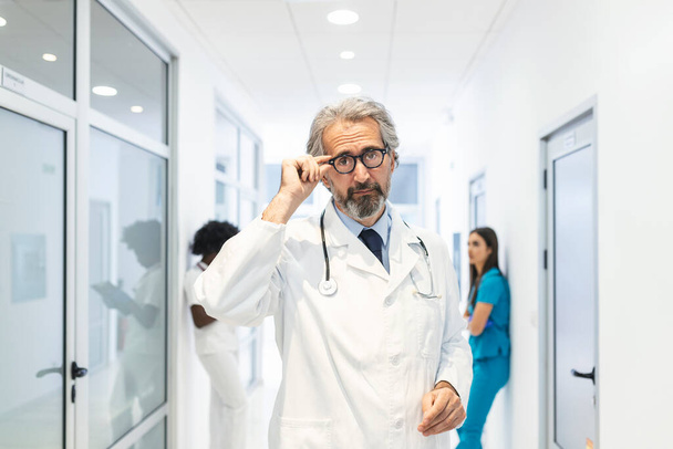Hastane koridorunda gözlüğünü tutan kendinden emin olgun bir doktorun portresi. Hastanede laboratuvar önlüğü giyiyor.. - Fotoğraf, Görsel