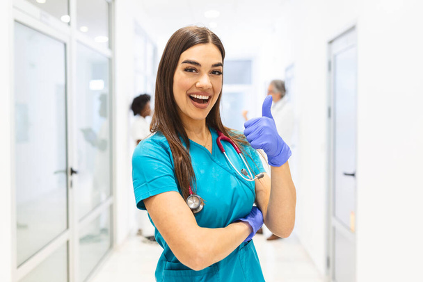 Портрет Усміхненого жіночого лікаря, який носить чагарники у багаторазовому лікарняному коридорі з пальцями вгору. - Фото, зображення