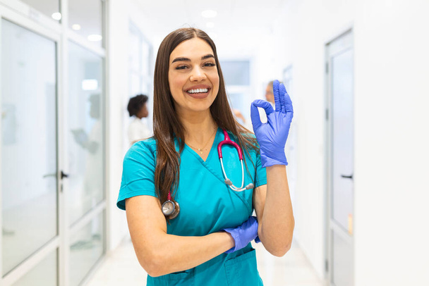 Junge Ärztin, die im Krankenhaus arbeitet und medizinisches Personal, sie zeigt OK-Zeichen. Krankenschwester steht auf dem Gehweg und lächelt mit verschränkten Armen. Sie trägt Peelings und ein Stethoskop. - Foto, Bild