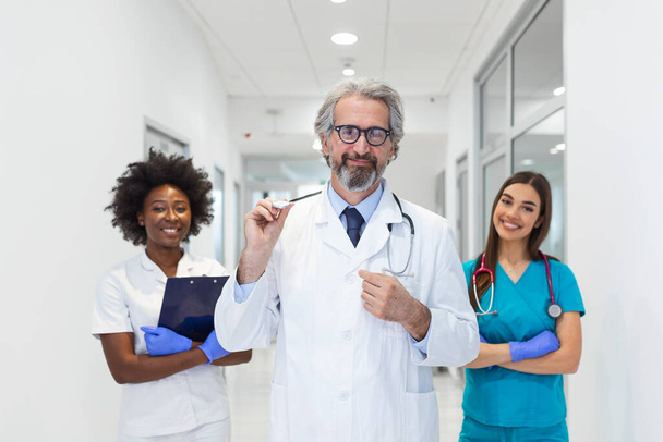 A multi-ethnic group of medical staff . They are dressed in medical scrubs and white lab coats with stethoscopes around their necks. Some standing in the back. The focus is on an mature man in front - Fotoğraf, Görsel