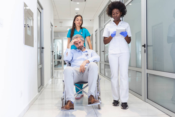 Senior man patient in wheelchair sitting in hospital corridor with African American female doctor and nurse. African American doctor and nurse in scrubs with patient in hospital wheelchair in corridor of specialist care clinic - Foto, afbeelding