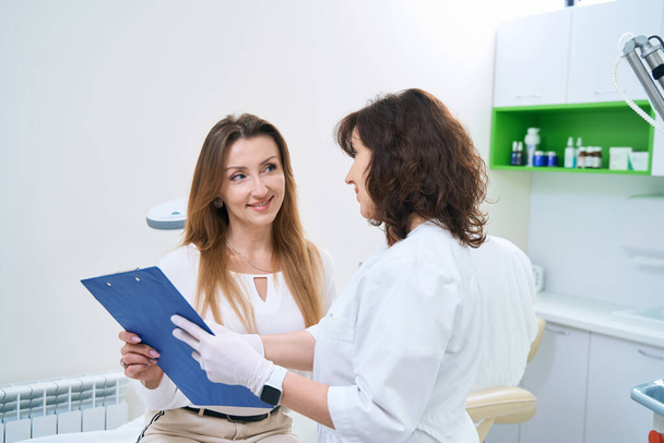 Smiling female patient of clinic with doctor examining medical record - Photo, image