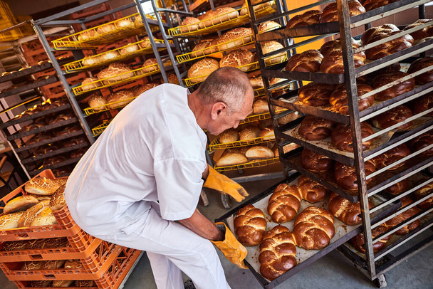 Baker sostiene una bandeja con pan caliente fresco en sus manos contra el fondo de los estantes con pasteles en la panadería. Producción industrial de pan - Foto, Imagen